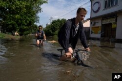 Местные жители Новой Каховки пытаются проехать на велосипедах по затопленной дороге после подрыва плотины Каховской ГЭС в ночь на 6 июня 2023 года